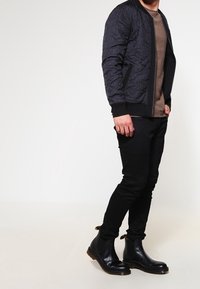 Black quilted bomber jacket with textured fabric, paired with a brown shirt and black fitted pants, and black ankle boots.