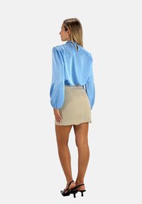 Blusa di raso azzurro chiaro con maniche a palloncino, dettaglio a chiave sulla schiena, abbinata a una minigonna beige aderente e sandali neri con cinturini.