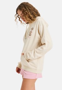 Crème hoodie met een voorzak, trekkoord capuchon en bloemenontwerpen in bruin. Gecombineerd met roze shorts. Zachte stof en een ontspannen pasvorm.