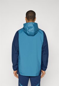 Lichtgewicht capuchonjack met een bicolor ontwerp in blauw en marineblauw, voorzien van elastische manchetten en een ontspannen pasvorm. Textuurstof met een gladde afwerking.