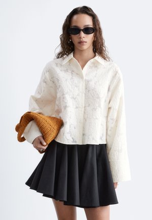 Femme aux longs cheveux bouclés portant des lunettes de soleil ovales noires, une chemise blanche texturée à boutons, une jupe plissée noire, tenant un vêtement tricoté brun plié.