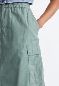 Zielona spódnica do kolan wykonana z lekkiego materiału, z elastycznym pasem i boczną kieszenią. Gładka faktura z minimalną liczbą szwów.