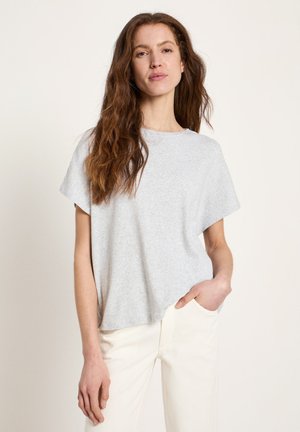 Woman with long brown hair wearing a light gray short-sleeve shirt and white pants, one hand in pocket, standing against plain light background.