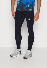 Leggings atléticos negros con un ajuste ceñido, que cuentan con el logo de New Balance en blanco, combinados con zapatillas de running grises con acentos en neón.