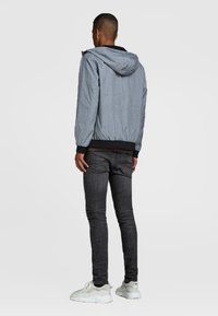 Chaqueta ligera y gris con capucha, puños y dobladillo elásticos, combinada con jeans oscuros y zapatillas blancas. La textura parece suave.