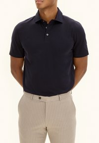 Navy blue short-sleeve polo shirt made of smooth fabric, featuring a classic collar and three-button placket, paired with light striped trousers.