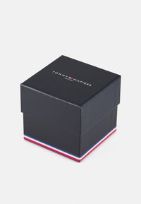Scatola nera con coperchio, caratterizzata dal logo "TOMMY HILFIGER" in bianco. La base ha un accento a strisce rosse, bianche e blu. Forma quadrata, texture liscia.
