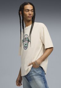 Beige bomulds t-shirt med et farverigt grafisk design, der viser en sol og et øje, parret med lyseblå denimjeans.