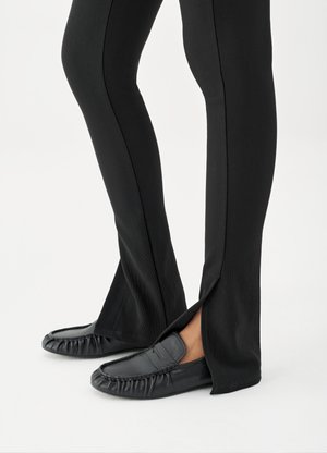 Jalat, joissa mustat levenevät housut ja sivuhalkiot, yhdistettyinä mustiin nahkajalkineisiin, valkoisella taustalla.