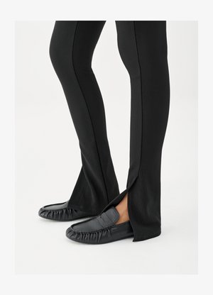 Jalat, joissa mustat levenevät housut ja sivuhalkiot, yhdistettyinä mustiin nahkajalkineisiin, valkoisella taustalla.