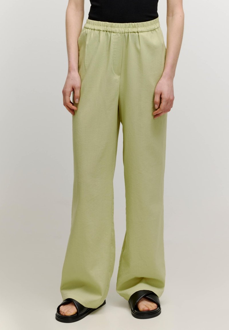 Pantalons large vert clair en tissu doux avec une taille élastique et deux poches latérales, associés à des sandales plates noires.