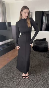 Svart klänning med långa ärmar och figurnära design, hög krage och detalj med söm mitt fram. Bärs med svarta remsandaler med klack. Gles bakgrund.