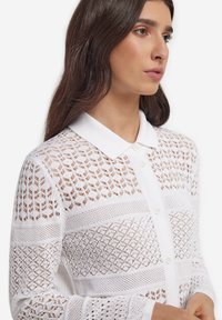 Biały dziergany kardigan z kołnierzem, z mieszanką wzorów ażurowych i gładkich sekcji. Zapięcie na guziki i detale z teksturowanej tkaniny.