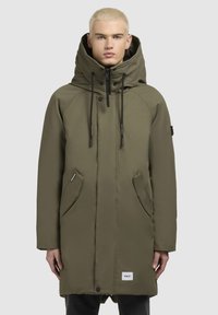 Olivengrøn parka-jakke med høj krave, justerbar hætte, forlommer og lynlåslukning. Har sorte snore og logo-patch.