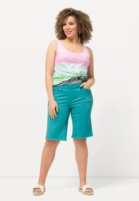 Canotta leggera a righe nei toni del rosa, verde e nero. Pantaloni corti teal, lunghi fino al ginocchio, con orlo sfilacciato. Modella che indossa sandali bianchi.