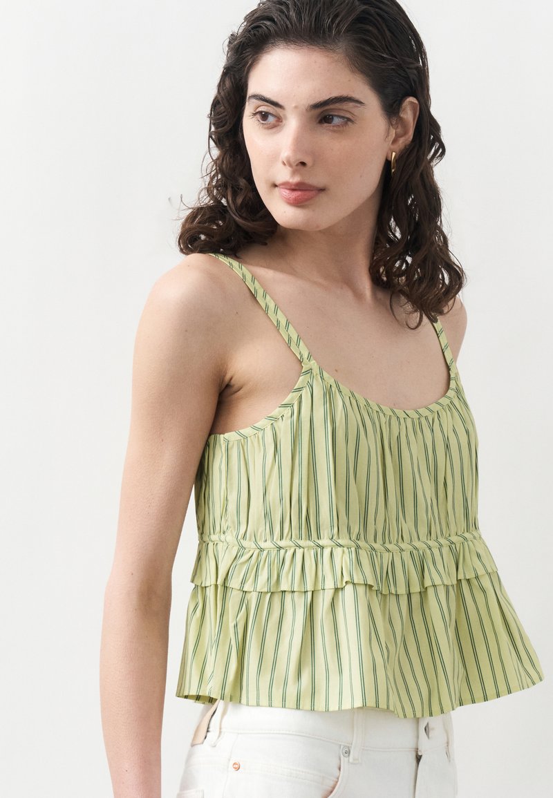 Femme aux cheveux bouclés et foncés, portant un haut sans manches vert clair rayé à volants et un pantalon blanc, regardant vers la gauche sur un fond uni.