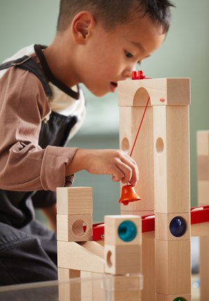 Holzbauklötze in natürlicher Ausführung mit bunten, runden Akzenten. Ein Kind spielt mit einer roten Glocke, die an einer Blockstruktur mit einer Schnur befestigt ist.