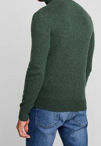 Stickad grön tröja med hög krage och ribbade ärmslut, kombinerad med blåjeans. Mjuk textur med en figurnära silhuett.