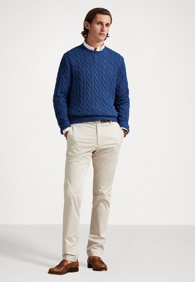 Μπλε ζακάρ πουλόβερ, μπεζ παντελόνια και καφέ δερμάτινα υποδήματα. Το πουλόβερ διαθέτει υφασμάτινο σχέδιο, ενώ τα παντελόνια είναι λεία και στενά.