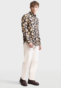 Blumenhemd in Schwarz mit orangefarbenen und weißen Blumen, kombiniert mit weißen Hosen und braunen Schnürschuhen. Texturiertes Material, lässige Passform.