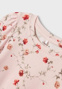 Chemise en coton rose avec des manches bouffantes, motif floral avec des roses rouges et des feuilles vertes, encolure ronde et une petite étiquette de marque.