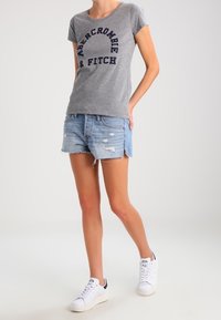 Persona con t-shirt grigia Abercrombie & Fitch, shorts di denim strappati e sneakers bianche, in piedi con una mano in tasca.