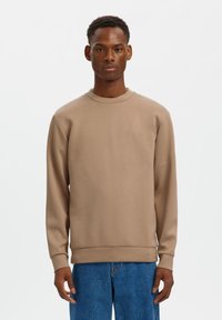 Καφέ φούτερ με στρογγυλό λαιμό και μακριά μανίκια, κατασκευασμένο από ομαλό ύφασμα. Διαθέτει ριμπ μανσέτες και τελείωμα, φορεμένο με μπλε τζιν.