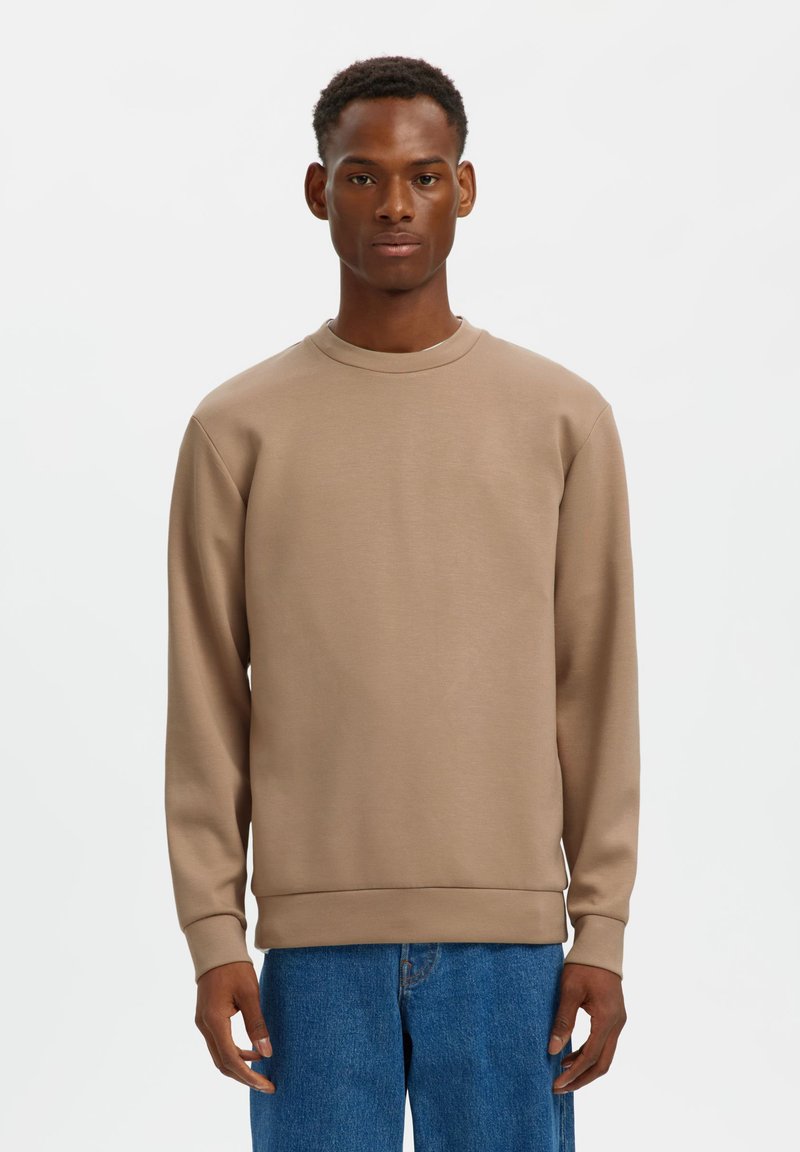 Καφέ φούτερ με στρογγυλό λαιμό και μακριά μανίκια, κατασκευασμένο από ομαλό ύφασμα. Διαθέτει ριμπ μανσέτες και τελείωμα, φορεμένο με μπλε τζιν.