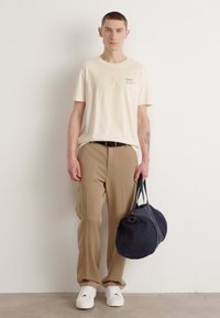 Lichtbeige T-shirt met logo, tan broek, witte sneakers en een donkerblauwe sporttas. Model staat tegen een effen achtergrond.