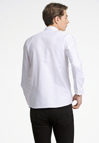 Camisa blanca de manga larga con puños abotonados, que presenta un diseño limpio y una textura suave, combinada con jeans negros. Vista desde la parte posterior.
