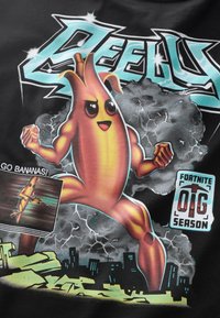 Sort T-shirt med en tegneserie-banan-superhelt med muskuløse arme, orange-gul krop og udtryksfulde øjne, omgivet af en stormfuld baggrund.