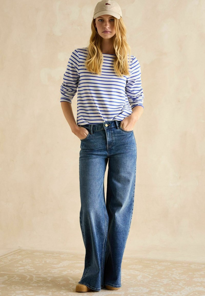 Sinine ja valge triibuline pikkade varrukate särg, millele on sobitatud kõrge vöökoht ja laiad sinised teksapüksid ning beež müts; vabalt langev, mugav stiil.