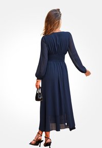 Granatowa długa sukienka z przezroczystymi długimi rękawami i marszczoną talią, z falbanistą spódnicą, noszona z czarnymi sandałami na obcasie i małą torebką.