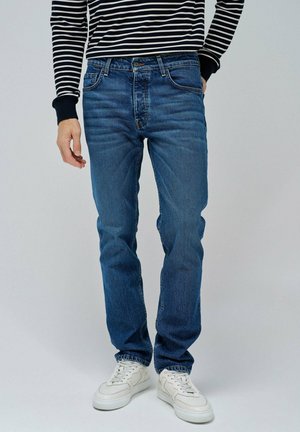 Jeans de mezclilla azul con un corte recto, diseño de cinco bolsillos y un ligero desvanecimiento. Llevados con una camisa de manga larga a rayas en blanco y negro y zapatillas blancas.