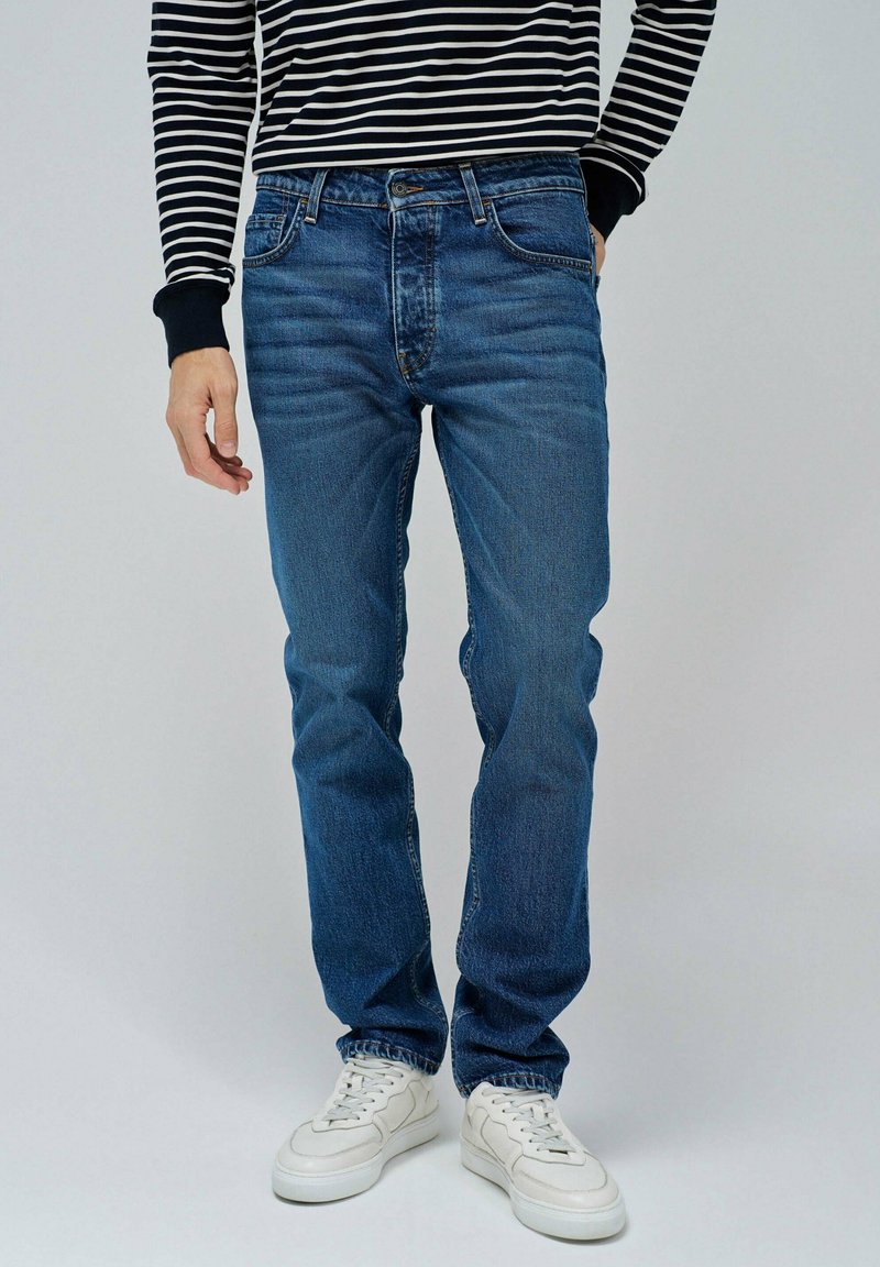 Jeans de mezclilla azul con un corte recto, diseño de cinco bolsillos y un ligero desvanecimiento. Llevados con una camisa de manga larga a rayas en blanco y negro y zapatillas blancas.
