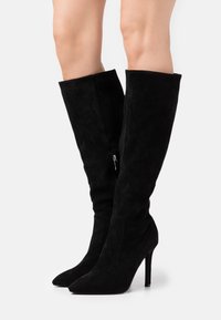 Botas negras por encima de la rodilla hechas de ante, con punta afilada, tacón stiletto alto y detalle de cremallera lateral. Textura suave y diseño elegante.