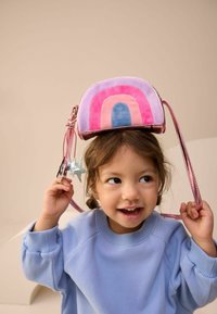 Torebka w tęczowym wzorze z różowego, fioletowego i niebieskiego aksamitu; ma metalowe różowe paski i srebrny urok w kształcie gwiazdki. Dziecko trzyma torbę na głowie.