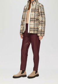 Osoba v béžovém, černém a broskvovém kostkovaném saku, vínových kožených kalhotách, světle hnědých semišových botách a světle béžovém topu na jednobarevném pozadí.