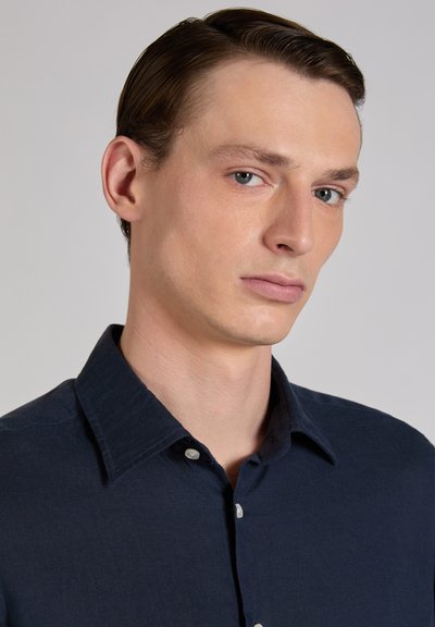 Joven con piel clara y ojos azul claro, vistiendo una camisa abotonada de color azul marino oscuro, mirando ligeramente hacia un lado sobre un fondo liso.