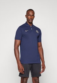 Marineblauwe poloshirt met een drieknoopsluiting, korte mouwen, Nike-logo in goud en een ronde patch met een teamembleem.