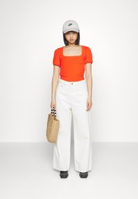 Top curto de manga curta riscado em vermelho, jeans largos brancos, calçado preto, boné cinza da Nike e uma mala trançada com alças de contas.