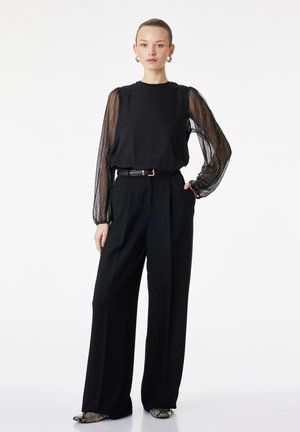 Jeune femme debout portant un haut noir à manches longues transparentes à pois, un pantalon noir taille haute à jambes larges, une ceinture, des boucles d'oreilles créoles dorées et des chaussures en peau de serpent.
