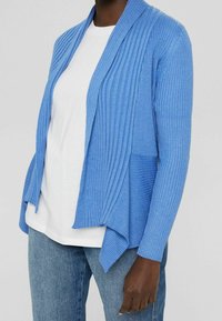 En person som bär en ljusblå ribbad kofta över en vit skjorta och blå jeans, som står mot en enkel ljus bakgrund.