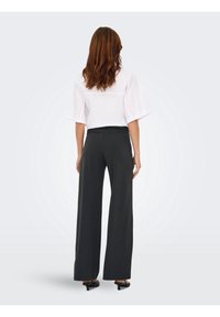 Femme aux longs cheveux bruns, debout de dos à la caméra, portant un chemisier blanc à manches courtes, un pantalon noir à jambes larges et des talons noirs.