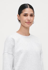 Jovem mulher com cabelo liso e escuro preso, brincos de argola prateados, vestindo um sweatshirt cinza claro, sorrindo suavemente contra um fundo branco simples.