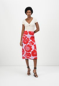 Cremeöverdel med en knuten detalj, kombinerad med en lavendelfärgad kjol med stora röda blommönster. Modellen bär svarta sandaler med remmar.