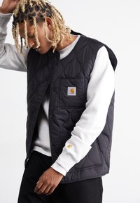 Black quilted sleeveless vest with snap buttons and a front pocket, worn over a grey sweatshirt; features a yellow logo on the pocket and cuff.