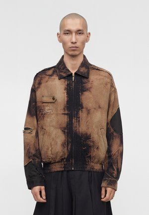 Άνδρας μοντέλο με ξυρισμένο κεφάλι που φοράει καφέ και μαύρο καρό μπουφάν με φερμουάρ, με φθαρμένες λεπτομέρειες και μαύρα έμπλαστρα στους αγκώνες.