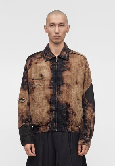 Modèle masculin à tête rasée portant une veste zippée à carreaux marron et noir avec des détails usés et des coudières noires.