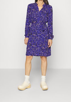 Femme portant une robe portefeuille fleurie violette mi-longue à manches longues, des chaussettes blanches et des sabots en bois beige.